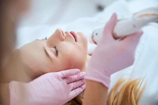 Blonde woman receiving the fractional microneedling treatment Cropped photo of a doctor in latex gloves directing the beauty device at the patient face